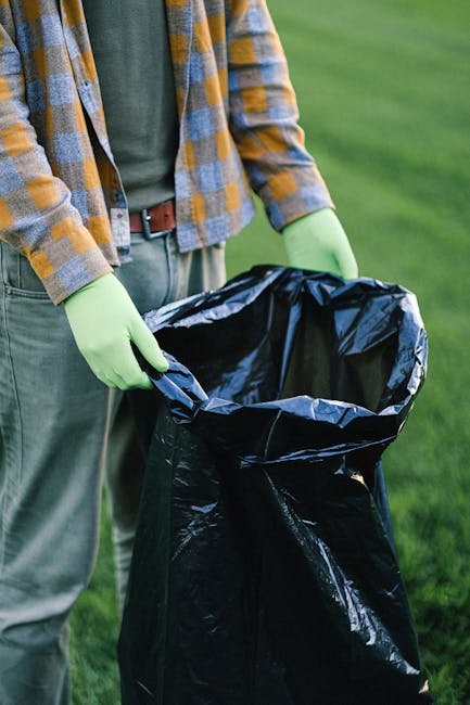 The image depicts a person outdoors in a grassy area, dressed in casual clothing including a plaid shirt with shades of yellow, grey, and blue, and green rubber gloves. They are holding open a large black rubbish bag made of durable plastic, which is partly expanded to show its interior. The surface of the bag is shiny and smooth, with some crumpling visible around the edges. The person's waist and hands are visible, with the bag positioned close to the ground, suggesting they are in the process of collecting waste or preparing for rubbish disposal. The background features a well-maintained green lawn, indicating an outdoor setting suitable for waste collection or cleanup activities. This scene subtly reflects an independent or private approach to rubbish management, aligning with the theme of non-local-authority waste handling and on-site rubbish collection services. The lighting appears natural, with soft shadows suggesting daylight conditions, and the overall image maintains a neutral and professional tone suitable for illustrating rubbish removal services.