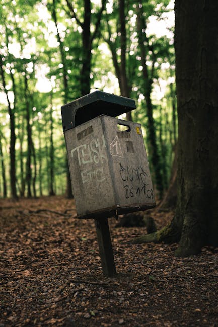A grey metal public mailbox with a black lid, situated in a wooded area with tall trees and green foliage in the background. The mailbox appears weathered, with visible graffiti and tags on its surface. It is mounted on a slanted metal post embedded into the ground, which is covered with brown, textured soil and scattered small stones. The surrounding environment is a natural setting, possibly part of a park or forest trail, with dappled sunlight filtering through the leaves. The mailbox’s design is standard for street or park use, but its placement among trees and natural terrain suggests it may be part of an urban area that provides alternative or independent waste or document collection services, such as a community notice point or a site for discarded small items. The scene emphasizes a contrast between the functional object and the natural environment, highlighting the importance of responsible waste handling in outdoor spaces, with occasional reference to waste disposal or collection activities relevant to local rubbish removal services.