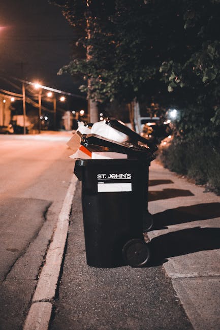 A black wheeled rubbish bin positioned on the edge of a residential sidewalk at night. The bin is labeled with the name 'St. John's' and is filled with a variety of waste materials, including cardboard boxes and plastic items, some of which are overflowing and leaning against the exterior. The surrounding environment features a dimly lit street with orange-hued streetlights illuminating the scene. In the background, there are trees with dark foliage and street furniture, such as poles and parked vehicles, visible further along the road. The pavement appears clean and well-maintained, and the bin's presence suggests an alternative waste management solution, possibly a private rubbish collection service, in contrast to council-provided refuse disposal. The scene's lighting and setting emphasize a typical household or community waste disposal point, with the focus on the waste container prepared for pickup, aligning with the context of rubbish removal services near Tufnell Park.