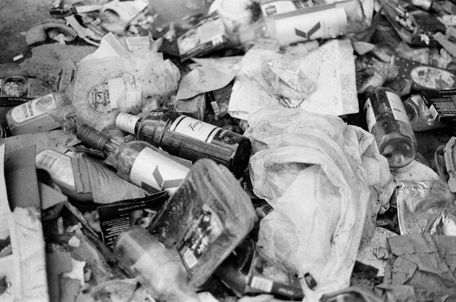 A large collection of mixed household waste and rubbish is piled up on a paved urban street near a parking area. The refuse includes cardboard boxes, plastic bags, paper, and packaging materials, with some items spilling onto the surrounding pavement. In the foreground, several brown cardboard boxes and plastic containers are visible, along with crumpled papers and miscellaneous debris. Several black and red rubbish bags are stacked or scattered among the waste, some partially opened revealing rubbish inside. Behind the pile, a medium-sized, grey metal bin marked for mixed paper and cardboard stands overflowing with packaging and paper waste. To the left, a parked grey car with a British registration plate is adjacent to a low metal railing separating the waste from the parking area. In the background, a commercial building with signage and a shopfront is visible, with a blue metal scaffold structure installed on the façade. The scene is captured during daylight hours, with natural lighting illuminating the urban environment. This image reflects typical on-site waste accumulation prioritising private rubbish disposal over municipal collection, suitable for professional rubbish removal services like those offered by [COMPANY_NAME].