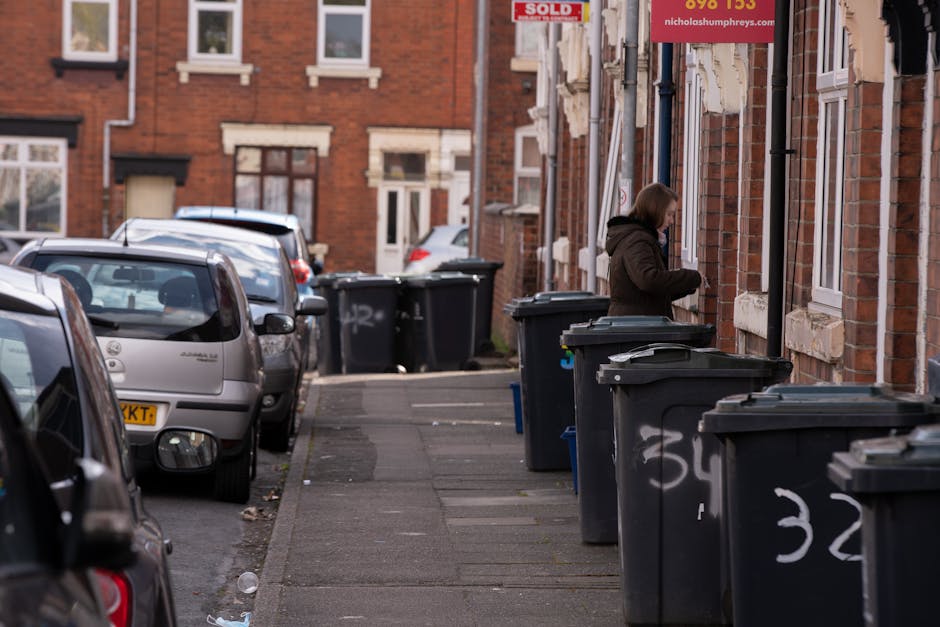 A residential street scene featuring multiple black wheeled rubbish bins with closed lids lined up along the pavement outside brick terraced houses. The bins are positioned in front of the houses, which have light-colored window frames and brick facades, with some displaying small signage or advertisements. A woman dressed in a dark coat is seen lifting or sorting items into one of the bins, indicating active waste disposal or collection activity. Parked cars, including a silver hatchback and other vehicles, line the street on the left side of the image, with some showing visible registration plates. The environment appears to be in an urban area with a slightly overcast or cloudy sky, casting diffuse natural light across the scene. The setting suggests a typical neighbourhood where private waste collection services, like those from rubbishcollectiontufnellpark.co.uk, are used to manage household rubbish, with the bins arranged for collection or on-site disposal. The overall composition reflects everyday waste management practices in a densely populated residential area, emphasizing the role of independent rubbish removal in urban waste handling.
