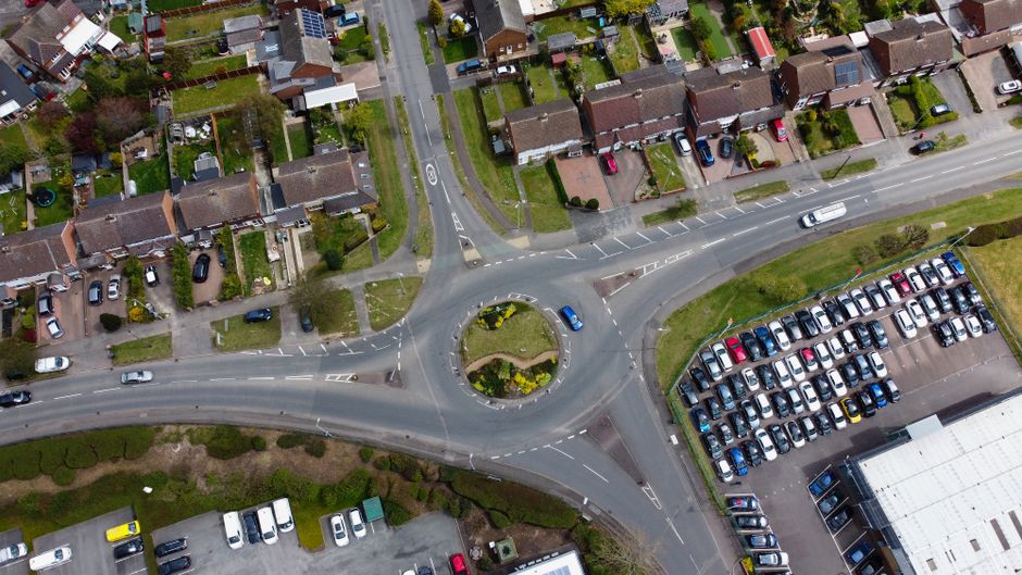 An aerial view of a roundabout located within a residential area, featuring a central landscaped island with small shrubs and yellow flowering plants. Surrounding the roundabout are several lanes of asphalt road with white dashed and solid lines indicating traffic directions. To the left, a row of single-family houses with paved driveways, small front gardens, and diverse roof coverings and exterior finishes can be seen. Parked cars are visible along the streets and in driveways, with some on the pavement. On the right, there is a large parking lot with numerous vehicles arranged in neat rows, adjacent to a commercial building with a flat, white roof. The environment is well-lit by natural daylight, and the area appears clean and orderly, demonstrating typical urban traffic and parking features. This scene is indicative of private, independent waste collection opportunities, as seen in the context of rubbish removal services for local residential and commercial properties, such as those provided by [COMPANY_NAME].